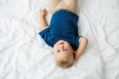 Mavi tişörtlü sarışın bir çocuk beyaz çarşaflı bir yatakta yatıyor. Bebek yatakta otururken oynar. Mavi gözlü küçük bir çocuk.