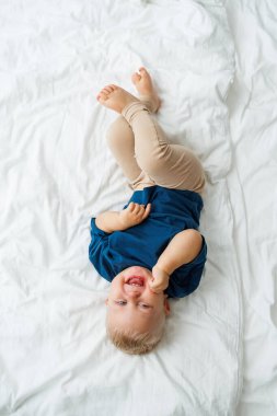 Mavi tişörtlü sarışın bir çocuk beyaz çarşaflı bir yatakta yatıyor. Bebek yatakta otururken oynar. Mavi gözlü küçük bir çocuk.