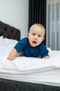 Mavi tişörtlü sarışın bir çocuk beyaz çarşaflı bir yatakta yatıyor. Bebek yatakta otururken oynar. Mavi gözlü küçük bir çocuk.