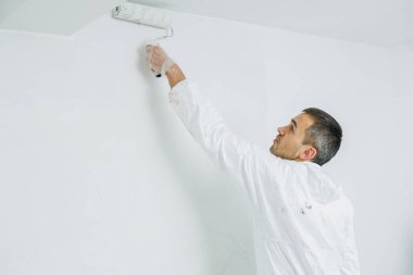 Ev tadilatı yapan adam. Endüstriyel konu. Duvarları boyuyorum. Beyaz tulumlu bir bina. Duvarı beyaz boya silindiriyle boyuyor..