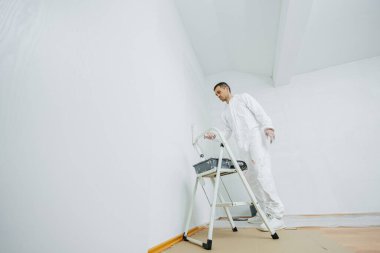 Binanın tadilatı. Siyah saçlı genç bir adam dairedeki duvarı beyaza boyuyor. Elinde bir silindir tutuyor. Ucuz tamirat kavramı.