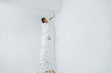 Elinde silindir olan beyaz üniformalı bir adam evde tavanı beyaza boyuyor. Binanın yenilenmesi.