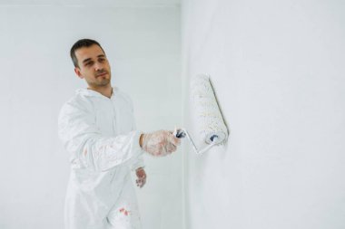 Elinde silindir olan beyaz üniformalı bir adam evde tavanı beyaza boyuyor. Binanın yenilenmesi.