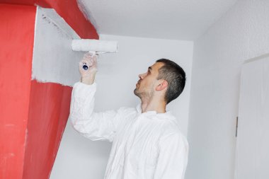 Beyaz tulumlu inşaat işçisi beyaz boya silindirli bir duvar boyuyor. Ev tadilatı. Endüstriyel konu. Duvarları boyuyorum.