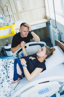 Bir adam, doğurmak üzere olan karısının elini tutar. Genç bir kadında doğumun başlangıcı. Doğum sırasındaki kocanın desteği. Doğum partneri. Doğum odası..