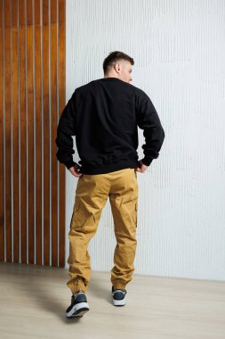 Gündelik kıyafet giymiş bir adam, kendine güvenen, benzersiz, desenli bir duvara karşı durarak, modern moda eğilimlerini ön plana çıkarıyor.