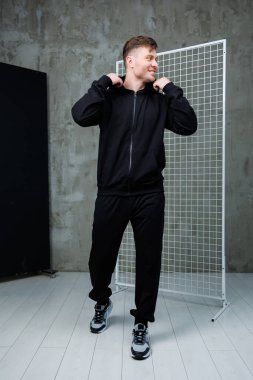 Genç bir adam, minimalist ve şık bir stüdyo ortamında modaya uygun siyah bir spor kıyafeti sergiliyor.