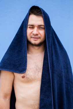 Üstsüz bir adam, mavi bir havluyla başının üstüne sarılı dururken kendine güvenen bir atmosfere sahiptir.