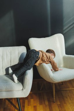 Neşeli bir çocuğun modern bir iç mekanda koltuklarda neşe içinde gezdiği ve dengede durduğu büyüleyici bir sahne.