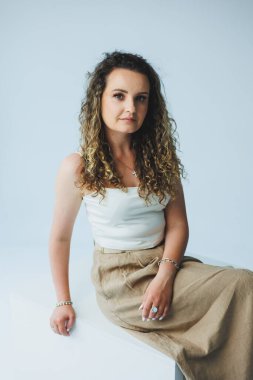 Sofistike bir kadın beyaz bir bluz ve altın bir etekle minimalist bir arkaplana karşı zarif bir şekilde poz verir..