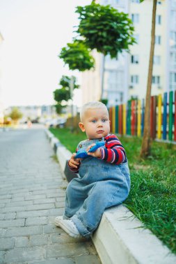 Sevimli bir çocuk kaldırımda oturuyor, neşeli ve canlı bir şehir ortamında güneşli bir günün tadını çıkarıyor.