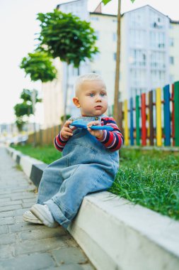 Neşeli bir çocuk kaldırımda oturur, etrafı renklerle çevrili, çocukluk mutluluğunun neşeli anlarını yakalar.