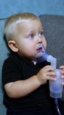 Küçük bir çocuk kanepede oturur ve astım spreyiyle nefes alır. Çocuklar için terapi amaçlı solunum. Çocuklar için solunum maskesi.