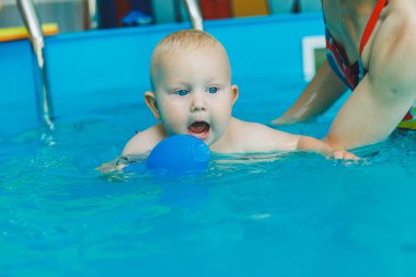 Bebek havuzda yüzüyor. Bir eğitmenin gözetiminde bebekler için yüzme havuzu. Havuzda çocuğu olan genç bir anne. Elinde bebekle suda eğlenen bir kadın.