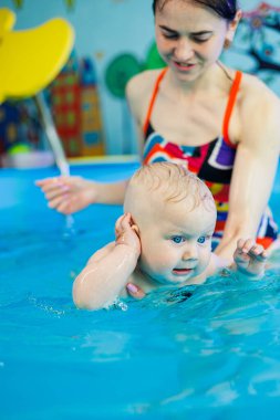 Bir antrenör havuzda bir bebekle ders veriyor. Çocuk antrenörle yüzmeyi öğrenir..