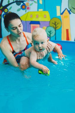 Bir eğitmenin gözetiminde yüzen bebekler için havuz. Havuzda çocuğu olan genç bir anne. Kadın kucağında bebek, suda eğleniyor.