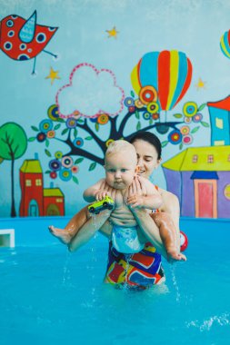 Bir eğitmenin gözetiminde yüzen bebekler için havuz. Havuzda çocuğu olan genç bir anne. Kadın kucağında bebek, suda eğleniyor.