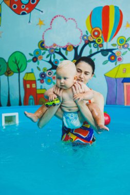 Bir eğitmenin gözetiminde yüzen bebekler için havuz. Havuzda çocuğu olan genç bir anne. Kadın kucağında bebek, suda eğleniyor.