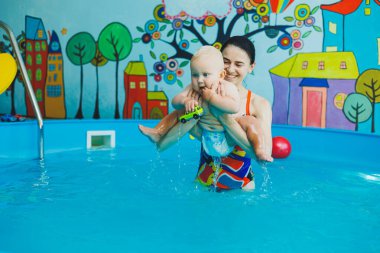 Bir eğitmenin gözetiminde yüzen bebekler için havuz. Havuzda çocuğu olan genç bir anne. Kadın kucağında bebek, suda eğleniyor.
