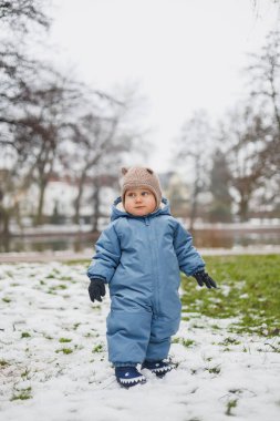 Ilık mavi bir kar elbisesi giymiş şirin küçük bir çocuk neşeyle güzel bir karlı arazide duruyor.