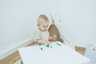 Bir yaşındaki küçük bir çocuk masada dururken plastik heykel yontuyor. Çocuk eğitici oyuncaklarla oynuyor..