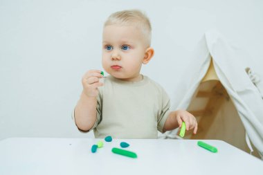 Bir yaşındaki küçük bir çocuk masada dururken plastik heykel yontuyor. Çocuk eğitici oyuncaklarla oynuyor..