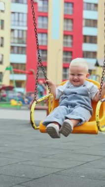 Küçük çocuk yazın güneşin altında oyun parkında oynuyor. Çocuk anaokulunun bahçesinde oynuyor. Renkli salıncakta hareketli bir çocuk. Çocuklar için sağlıklı bir yaz aktivitesi. Küçük çocuk sallanıyor.
