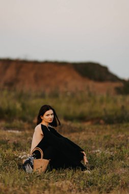 Gün batımında doğal bir manzarada oturan dalgalı siyah elbiseli bir kadının huzurlu görüntüsü huzuru çağrıştırıyor.