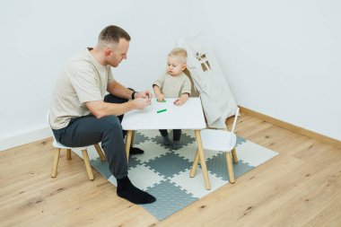 Sıcak bir ortamda, sevgi dolu bir baba meraklı bebeğiyle küçük bir masada oturup, hem kaynaşmayı hem de öğrenmeyi teşvik eden hoş ve yaratıcı bir anı paylaşır.
