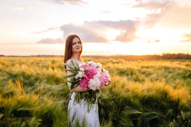Güzel bir gün batımı gökyüzünün altında, engin altın tarlaların arasında zarif bir buket tutan sakin bir gelin.