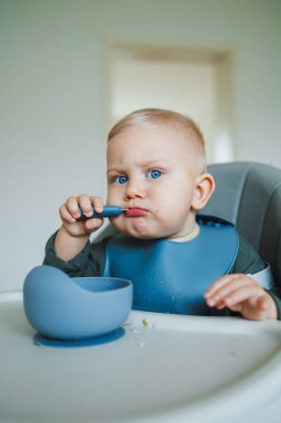 Işıl ışıl mavi gözleri ve dağınık bir ağzı olan büyüleyici bir bebek, parlak bir mutfak arka planına karşı kurulmuş, yüksek sandalyedeki kaşıkla kendilerini neşeyle besler.