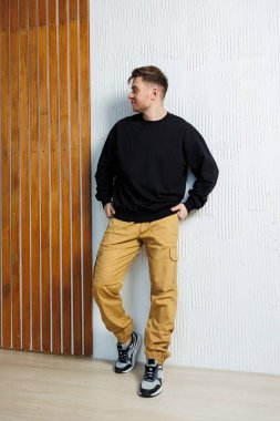 Bir adam rastgele duvara yaslanıyor, şık siyah bir kazak giyiyor ve göze çarpan sarı bir pantolon giyiyor.