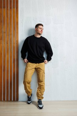 Siyah eşofman üstü ve sarı pantolon giyen bir adam rahat bir duruş ve tavırla duvara yaslanıyor.