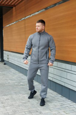 Şık, gri bir ceket ve rahat eşofman giyen bir adam modern bir binanın önünde kendinden emin bir şekilde duruyor.