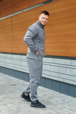 Eşofman altı ve rahat gri bir kazak giyen bir adam arkasında kırsal bir ahşap duvarın önünde duruyor.