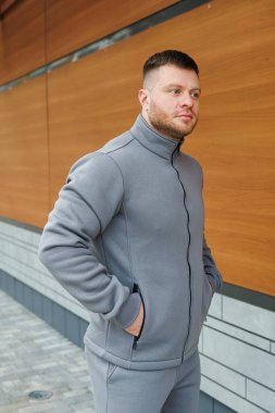 Şık, gri bir ceket giyen bir adam, ahşap bir duvarın önünde kendine güvenerek dikiliyor.