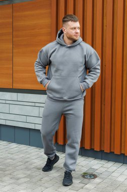 Gri kapüşonlu ve eşofmanlı bir adam sıradan bir şekilde ahşap duvarın önünde dikiliyor.