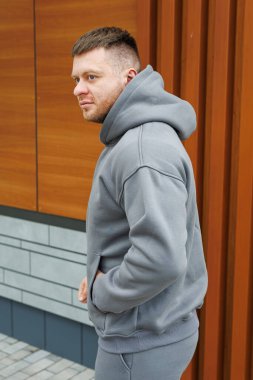 Gri kapüşonlu rahat giyinmiş bir adam şu anda kırsal bir ahşap duvarın önünde duruyor ve olay yerine derinlik katıyor.