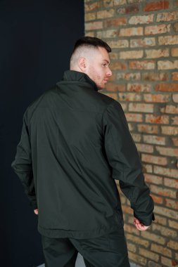 Parlak siyah ceketli bir adam arkasında uzanan süslenmemiş tuğla bir duvarın önünde duruyor.