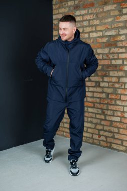 Mavi ceketli ve mavi pantolonlu bir adam, rustik görünümlü bir tuğla duvarın önünde güvenle duruyor.