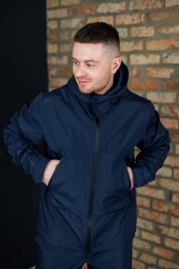 Mavi ceketli bir adam eski bir tuğla duvarın önünde duruyor. Zamanla yıpranmış ve yaş ve karakter belirtileri gösteriyor.