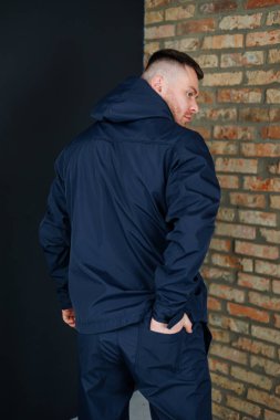 Mavi ceketli bir adam, aşınma ve yaşlanma belirtileri gösteren eski bir tuğla duvarın önünde güvenle duruyor.