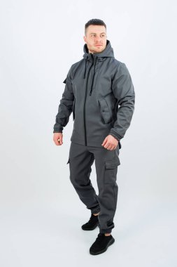 Şık gri bir ceket ve uyumlu pantolon giyen bir adam beyaz bir arka plana karşı belirgin bir şekilde duruyor ve kıyafetini güzel bir şekilde gösteriyor.