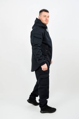 Tarz sahibi siyah ceketli ve siyah pantolonlu bir adamın parlak beyaz bir arkaplanda yürürken görülmesi çarpıcı bir kontrast oluşturur.