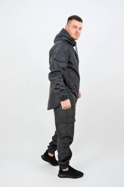 Sade beyaz bir arka planda duran bir adam şık bir siyah ceket giyiyor ve kıyafetini tamamlayan siyah bir pantolon giyiyor.