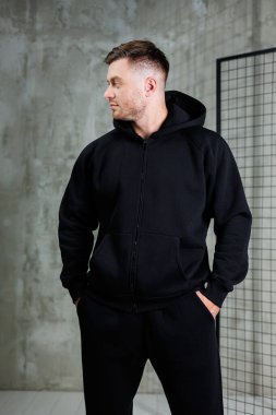 Siyah kapüşonlu ve siyah eşofmanlı bir adam elleri cebinde rahat bir şekilde duruyor.