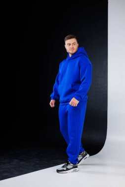 Mavi kapüşonlu ve mavi eşofmanlı bir adam belirgin siyah beyaz bir duvarın önünde duruyor.
