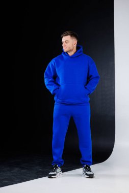 Mavi kapüşonlu ve mavi eşofmanlı bir adam siyah beyaz bir arka planın önünde güvenle duruyor.