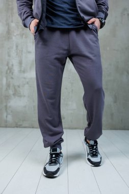 Şık bir ceket ve rahat bir eşofman giyen bir adam temiz, parlak beyaz bir zeminde güvenle duruyor.