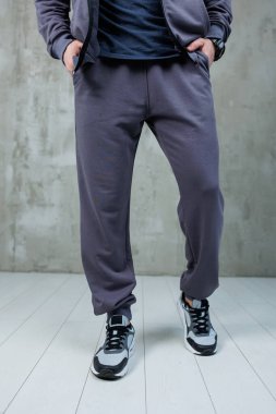 Şık bir ceket ve rahat bir eşofman giyen bir adam temiz, parlak beyaz bir zeminde güvenle duruyor.